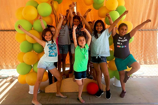 Group of happy teen girls posing and raising two hands in a green and yellow balloon background at a Preschool & Daycare Serving Hesperia, CA