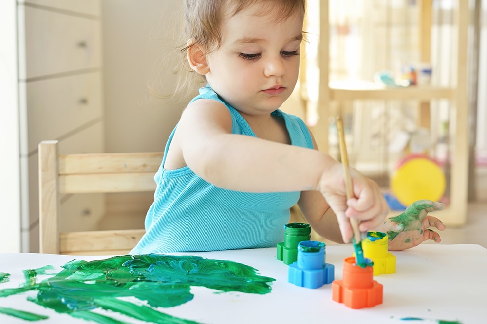 Cute little toddler child painting with paintbrush and colorful paints. Adorable baby girl drawing on white paper near window in light room at a Preschool & Daycare Serving Hesperia, CA