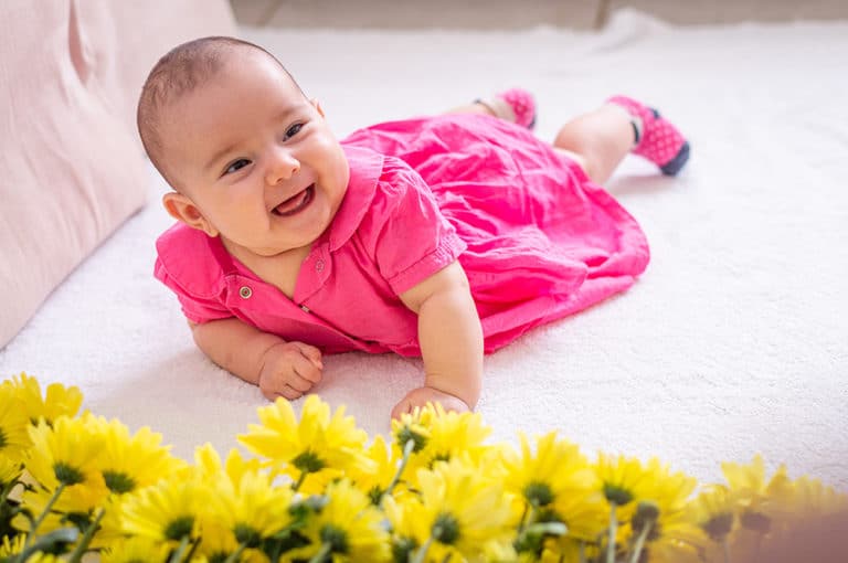 Baby in pink dress on white bed, hands resting next to yellow flowers laughs funny and shows tongue at a Preschool & Daycare Serving Hesperia, CA