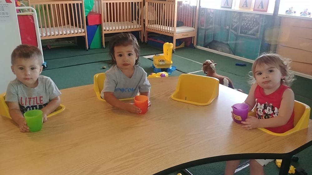 Group of toddlers drinking milk on a colorful plastic cups while sitting on a feeding table chair at a Preschool & Daycare Serving Hesperia, CA